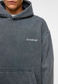 Grijze hoodie van een gestructureerde stof, met een vo zak en de tekst "CreateChange" in witte borduursel op de borst.