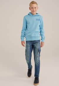 Vaaleansininen huppari, jossa on etupainatus, yhdistettynä sinisiin cargo-farkkuihin, joissa on sivutaskut ja tavallinen fit. Mustat kengät viimeistelevät asun.