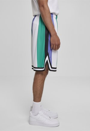 Hombre de pie en vista lateral que lleva pantalones cortos de baloncesto a rayas blancas, verdes, azules y negras, con calcetas blancas y zapatillas blancas.