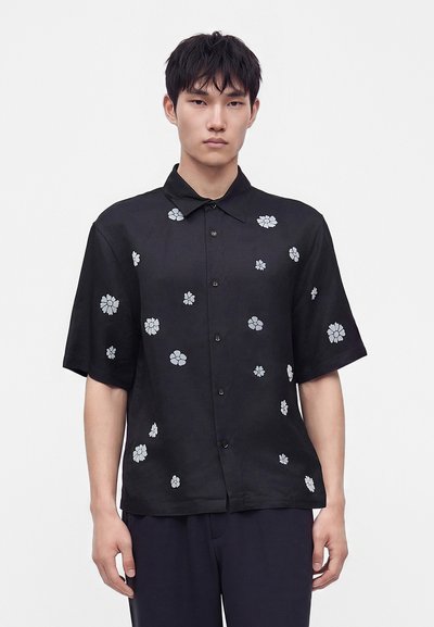Jeune homme portant une chemise noire à manches courtes avec des motifs floraux blancs, debout devant un fond clair uni.