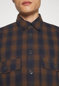 Rutigt flanellskjorta i marinblå och brun nyanser, knappstängning, krage och två framfickor med lock och knappar.