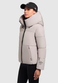 Helehall puffer-jakk, millel on kõrge krae, lukuga taskud ja mustad detailid. Omab tikandit ja silmatorkavat logo varrukal.