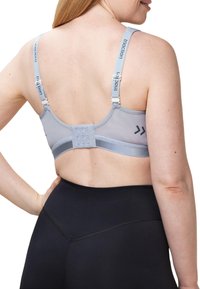 Lichtgrijze sportbeha met mesh-details en verstelbare banden; heeft een haak-en-oogsluiting aan de achterkant. Draagt met zwarte high-waisted leggings.