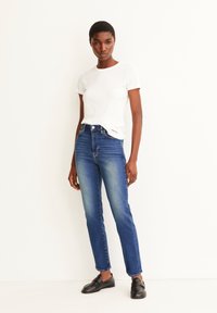 T-shirt blanc à manches courtes en côtelé associé à un jean en denim bleu clair taille haute. La mannequin porte des mocassins noirs et se tient devant un fond neutre.