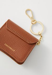 Portafoglio in pelle di colore beige con pattina, hardware dorato e portachiavi. Presenta il logo "Coccinelle" in rilievo. Forma rettangolare compatta.