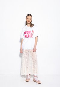 Biała koszulka z wyraźnym różowym napisem "ŚWIATOWY POKÓJ", nałożona na przezroczystą, kremową spódnicę, w zestawieniu z beżowymi butami i białymi skarpetkami.