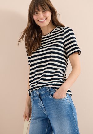 Donna sorridente che indossa una maglietta a maniche corte a righe bianche e nere e jeans blu, con una mano nella tasca dei jeans e sfondo chiaro.