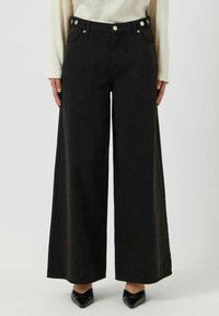 Svarta vida jeans med hög midja, featuring en knappselect, två framfickor och en slät textur. Bärs med spetsiga svarta skor.