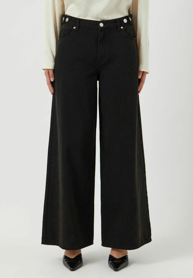 Svarta vida jeans med hög midja, featuring en knappselect, två framfickor och en slät textur. Bärs med spetsiga svarta skor.