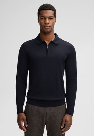 Polo a maniche lunghe blu marino con colletto zip, realizzato in un tessuto morbido, con orlo e polsini a coste. Indossato con pantaloni grigi texturizzati.