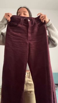 Person wearing a grey sweater holds up a pair of high-waisted, maroon corduroy pants in front of a plain wall.