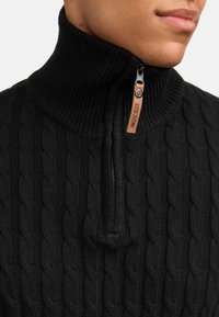 Schwarzer Pullover aus Kabelstrick mit hohem Kragen und halbem Reißverschluss. Mit einem Lederreißverschlussanhänger, auf dem der Text "INDICODE" steht.