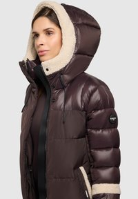 Giacca imbottita marrone con cappuccio beige soffice e dettagli coordinati. Presenta una chiusura con cerniera e bottoni, cappuccio rimovibile e tessuto testurizzato.