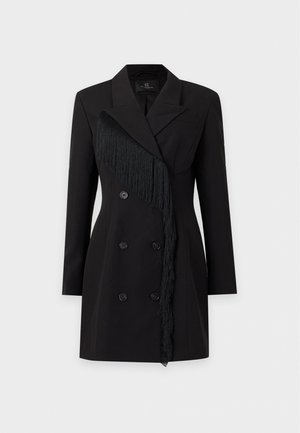 Un mannequin se tient devant un fond clair uni, vêtu d’une robe blazer noire croisée à franges, d’un collant transparent et de bottes montant jusqu’aux genoux.