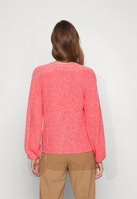 Strickpullover in lebhaftem Pink, mit langen, ballonförmigen Ärmeln, einem Rundhalsausschnitt und einer strukturierten Oberfläche. Trägt dazu beigefarbene Hosen.