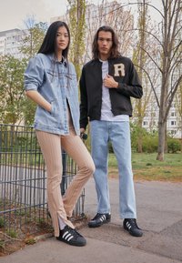 Džínová bunda, černý bomber s nášivkou „R“, béžové pruhované kalhoty, světle modré džíny. Oba mají na sobě černé tenisky Adidas. Zelené outdoorové pozadí.