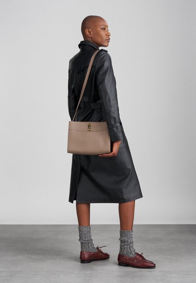 Beige Lederhandtasche mit glatter Textur, ausgestattet mit einem goldfarbenen Verschluss. Tragen von einem Model in einem schwarzen Ledermantel, grauen Socken und braunen Schuhen.
