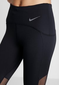 Czarne legginsy z wysokim stanem, wykonane z elastycznego materiału. Posiadają białe logo Nike oraz siateczkowe wstawki przy dolnej części nogawek.