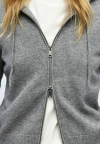 Zbliżenie na osobę ubraną w szary dzianinowy hoodie zapinany na zamek, noszącego biały T-shirt, z zamkiem częściowo rozpiętym w dolnej części.
