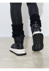 Bottines noires avec une surface texturée, une semelle intermédiaire blanche épaisse et des bordures en tricot noir. Semelle extérieure en caoutchouc nervurée pour une meilleure adhérence.