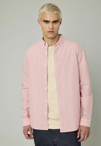 Chemise à rayures rouges et blanches avec un col classique, des manches longues et une coupe décontractée. Portée par-dessus un t-shirt beige.