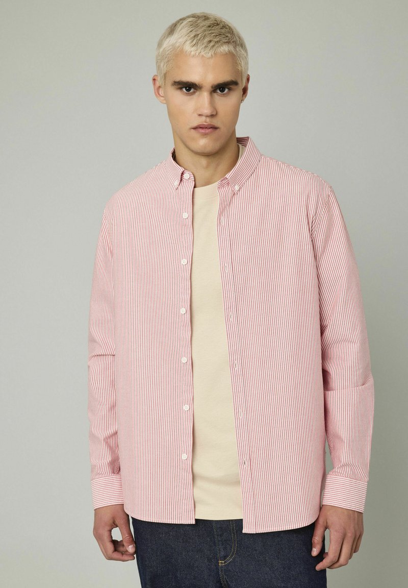 Chemise à rayures rouges et blanches avec un col classique, des manches longues et une coupe décontractée. Portée par-dessus un t-shirt beige.