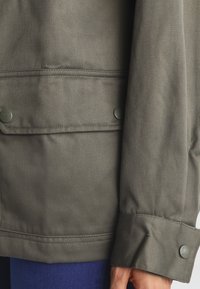 Veste vert olive avec une finition texturée, comportant deux poches avant et des fermetures à boutons-pression aux poignets.