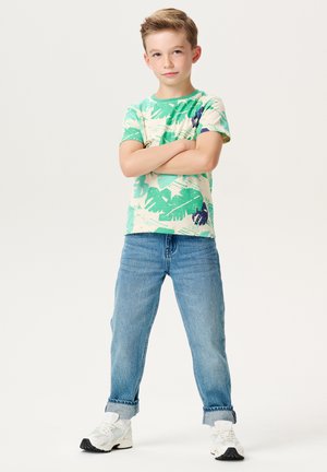 Grünes und cremefarben gemustertes T-Shirt mit abstrakten Formen, kombiniert mit hellblauen Jeans mit umgeschlagenen Ärmeln und weißen Sneakers mit schwarzen Akzenten.