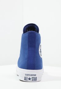 Blue canvas high-top sneaker with a white rubber sole and logo patch. Features a textured surface and rounded toe design.