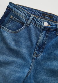 Dark blue denim jeans with a slightly faded texture, featuring a silver button and contrasting orange stitching. Notable front pocket design.