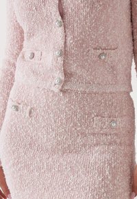 Bleg pink, tekstureret sæt bestående af en tætsiddende cardigan med perleknapper og en matchende nederdel med lommer og dekorative knapdetaljer.