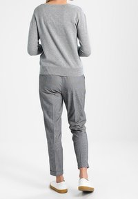 Person som står vänd bort med en grå tröja, mönstrade grå byxor och vita sneakers med bruna sulor på en vit bakgrund.