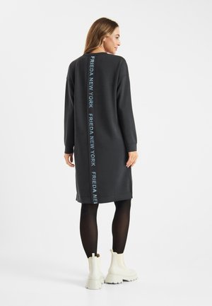 Zwarte sweatshirtjurk met een rechte snit, lange mouwen en achterop branding in lichtblauwe tekst. Draagt een zwarte panty en witte laarzen.