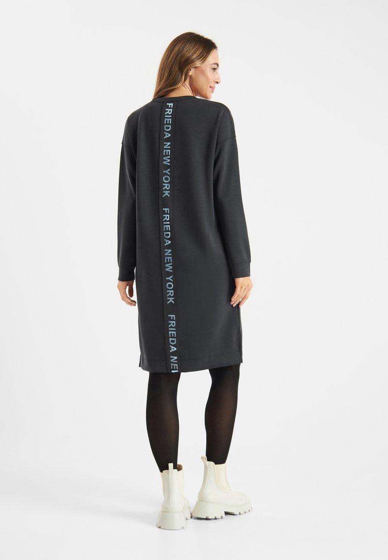 Zwarte sweatshirtjurk met een rechte snit, lange mouwen en achterop branding in lichtblauwe tekst. Draagt een zwarte panty en witte laarzen.