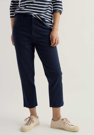 Persona con pantalones azul marino recortados, camisa a rayas azul marino y blanca, y zapatillas beige con cordones, de pie contra un fondo liso.