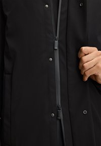 Czarna wodoodporna kurtka z zamkiem błyskawicznym z przodu, zatrzaskami i teksturowanym materiałem. Ręka widoczna trzymająca suwak.