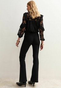 Femme aux cheveux blonds portant un chemisier floral noir transparent, un pantalon évasé noir, des chaussures à talons noires et des bracelets en or, se tenant devant un mur uni.