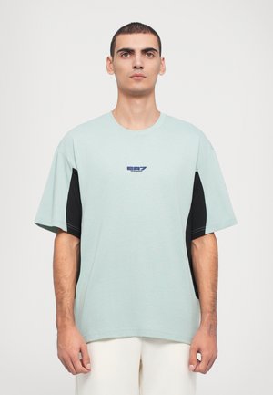 Tricou oversized de culoare verde mentă, cu panouri laterale negre, având un mic logo bleumarin pe piept și un guler rotund. Textură de țesătură netedă.
