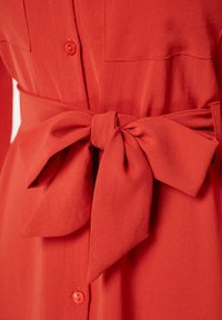 Rød stofkjole med knappelukning foran og bælte i taljen, der har en stor sløjfe. Materialet har en glat tekstur.