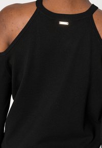 Pull noir à design épaules dénudées, encolure côtelée, avec une étiquette d'accent dorée à l'arrière. Texture en maille lisse, coupe ajustée.