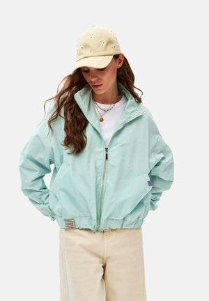 Žena v svetlomodrej bunde na zips, béžových nohaviciach, béžovej čiapke s kvetinovou výšivkou a viacerých náhrdelníkoch, pozerá sa dolu s rukami v kapsách.