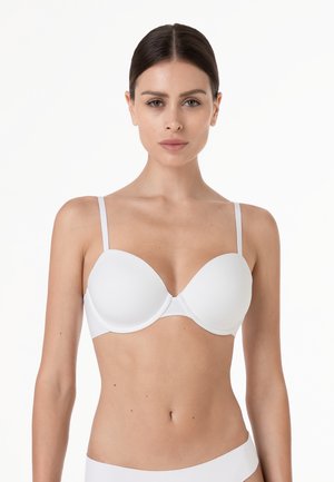 Donna con capelli scuri che indossa un reggiseno imbottito bianco, mostrata dalla vita in su su uno sfondo bianco uniforme.