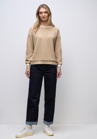 Sudadera beige de canalé con cuello redondo, ribete crema y un corte relajado. Combinada con jeans oscuros, remangados en los tobillos, y zapatillas blancas.