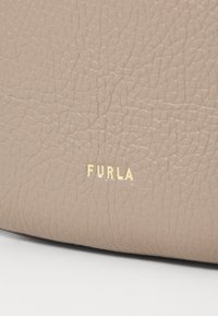 Bežová kožená plocha s textúrovaným povrchom, obsahujúca zlaté logo "FURLA". Materiál vyzerá mäkko s viditeľnými zrnitými vzormi.