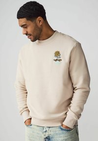 Beige collegepaita, jossa on ribattu kaula-aukko ja värikkäitä brodeerattuja kukka- sekä skeittimuotoiluelementtejä rinnassa. Pehmeä puuvillasekoitekangas.