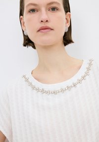 Top blanco texturizado con escote redondo, adornado con embellishments plateados que crean un patrón floral. La modelo lleva pendientes redondos y brillantes.