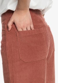 Pantalons en velours côtelé d'une couleur rose douce avec des rayures verticales, présentant une poche arrière et une main placée à l'intérieur de la poche.