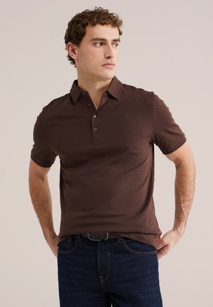 Muškarac u tamno smeđoj polo majici kratkih rukava i tamno plavim trapericama, gleda u stranu s rukama naslonjenim na bokove.