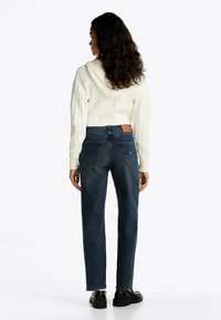 Sudadera blanca de punto trenzado, recortada y con capucha, combinada con jeans oscuros de corte recto que cuentan con dos bolsillos traseros y un parche de cuero.