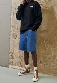 Musta huppari valkoisella logolla, siniset farkkushortsit, ruskeat korkeavartiset lenkkarit, valkoiset sukat; seisoo beigeä teksturoitua seinää vasten.